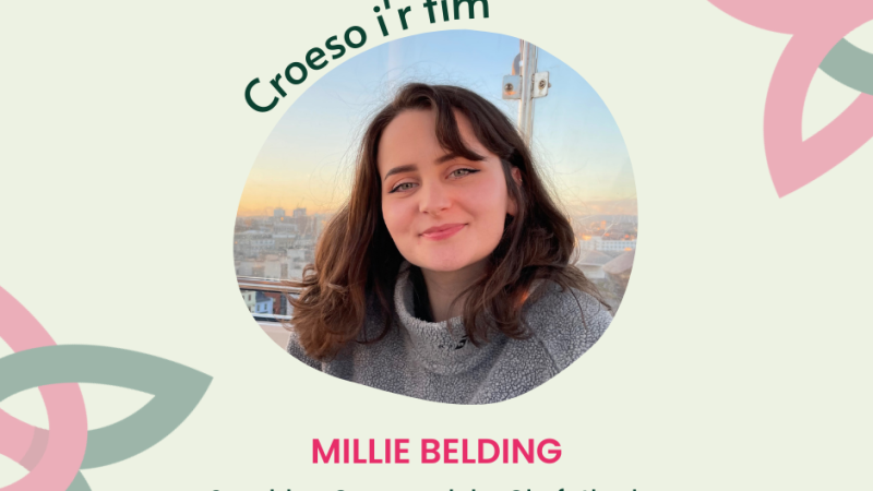 Image introduces a new team member to Fair Trade Wales' team with the company logo in the top left and the logo pattern in the background, which is a Dark green and pink celtic-like intertwined form on a cream coloured background. Photo in the middle is of Millie with a sunset in the background, she is a white female with brown hair. Text above the image reads 'croeso Millie' in dark green, with Millie's full name underneath in pink; Millie Belding and her job title; 'Swyddog Cymunedol a Chyfathrebu / Community and Communications Officer' in dark green.