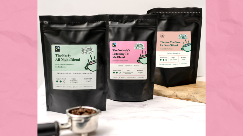 3 phecyn du o Goffi Tired Mums o'r chwith i'r dde: 'The party all night blend' gyda label lliw mint, 'the nobodys listening to me blend' gyda label pinc, a 'the are you sure it's decaf blend' mewn oren. Darllenwch fwy am ddisgrifiad pob cynnyrch ar eu gwefan https://tiredmumscoffee.co.uk/. Mae yna 'portafilter' gyda ffa coffi o fewn e, o flaen y bagiau coffi. Ac mae bwrdd torri wedi'i wneud o bren o dan y trydydd bag o coffi. Mae popeth wedi'i ddangos ar gownter marmor gwyn gyda wal wen y tu ôl iddo.