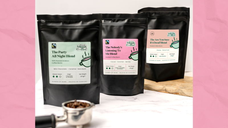 3 phecyn du o Goffi Tired Mums o'r chwith i'r dde: 'The party all night blend' gyda label lliw mint, 'the nobodys listening to me blend' gyda label pinc, a 'the are you sure it's decaf blend' mewn oren. Darllenwch fwy am ddisgrifiad pob cynnyrch ar eu gwefan https://tiredmumscoffee.co.uk/. Mae yna 'portafilter' gyda ffa coffi o fewn e, o flaen y bagiau coffi. Ac mae bwrdd torri wedi'i wneud o bren o dan y trydydd bag o coffi. Mae popeth wedi'i ddangos ar gownter marmor gwyn gyda wal wen y tu ôl iddo.